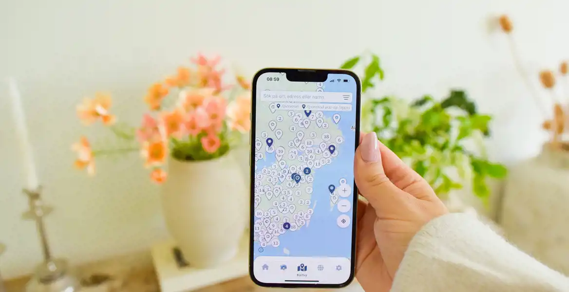 En hand håller en mobil med appen Loppinell öppen, där en karta över loppisar och second hand-butiker i södra Sverige visas. I bakgrunden står en vas med orange blommor och gröna växter på ett bord.