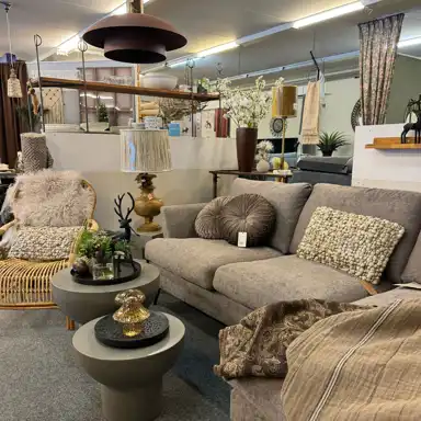 En hemtrevlig butiksmiljö med inredningsdetaljer och möbler i jordnära toner. I förgrunden syns en beige soffa med dekorativa kuddar och filtar, samt två runda soffbord med vaser, växter och prydnader. Till vänster står en fåtölj i rotting med ett mjukt fårskinn och en kudde. I bakgrunden syns lampor, gardiner, blommor och hyllor med inredningstillbehör.