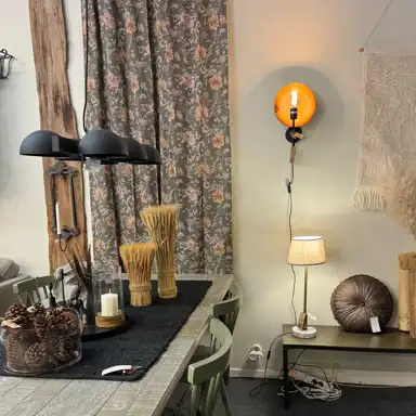 Ett mysigt inrett hörn av en butik eller ett hem, med ett rustikt träbord dekorerat med kottar, ljus och prydnadsax. På väggen hänger en varm orange vägglampa och en bordslampa med beige skärm står på ett sidobord tillsammans med en rund prydnadskudde. Blommiga gardiner i bakgrunden ramar in miljön och skapar en varm, lantlig känsla.