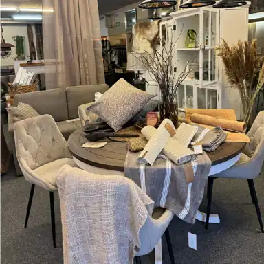 En inspirerande butiksutställning med ett runt träbord fyllt av textilier i jordnära toner, kuddar, löpare och rustika detaljer som träslevar och grenar i vas. Runt bordet står stoppade stolar i beige nyanser och i bakgrunden syns soffor, skåp och inredningsdetaljer i en varm och välkomnande miljö.