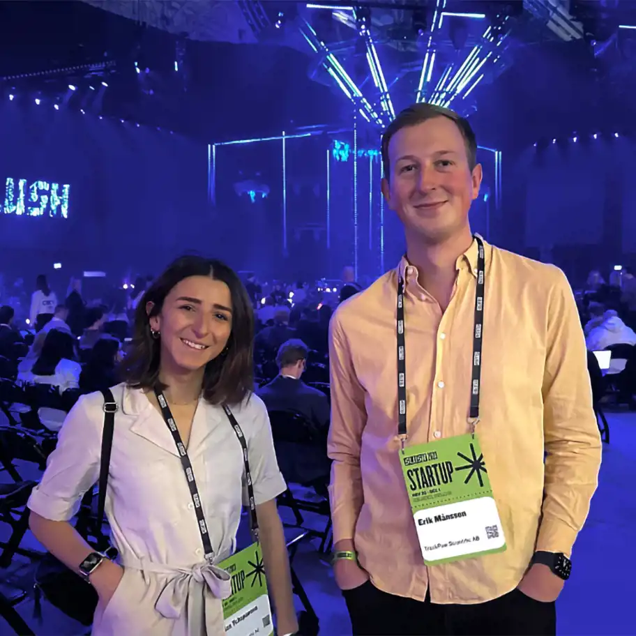 Två personer står och ler framför kameran på evenemanget Slush, omgivna av en stor publik i en mörklagd arena med blått och lila ljus. De bär ID-brickor som visar att de är startupdeltagare, varav en bricka visar namnet Erik Månsson och företaget TrackPaw Scientific AB. I bakgrunden syns scenljus och LED-strukturer som skapar en futuristisk stämning.