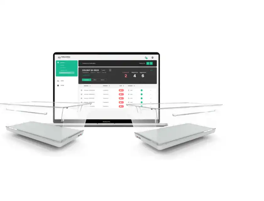 En dator med TrackPaw Scientifics mjukvara visas med två genomskinliga plastburar placerade framför. Skärmen visar en översikt av en koloni med ID-nummer, olika varningar och behandlingsdata. Gränssnittet är stilrent i svart, vitt och grönt och innehåller statistik som antal olösta ärenden, viktförändringar och aggressionsnivåer. Produkten används för att övervaka beteende och hälsa hos djur i forskningsmiljöer.